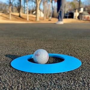 May include: A golf ball rests inside a bright blue circular target on a putting green. A golfer is in the background, preparing to putt. The scene is set outdoors on a sunny day.