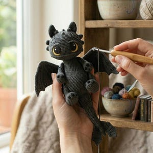 May include: A handmade, dark gray crochet dragon with large, expressive eyes and small wings. The dragon is being held in someone's hands, with a crochet hook visible. The background includes a wooden shelf with yarn and books.
