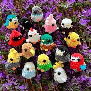May include: A collection of colorful, handmade crochet bird figurines. Each bird has a unique color scheme, including blue, yellow, red, and green. The birds are small, with detailed features like beaks and wings. They are arranged on a bed of purple flowers.
