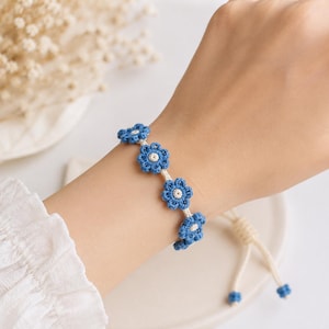 May include: A blue crochet flower bracelet with white accents. The bracelet features five blue flower designs connected by a white cord. The bracelet is adjustable with a drawstring closure.