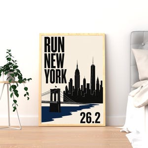 Peut inclure: Une affiche verticale encadrée présentant une silhouette noire de la ligne d'horizon de New York et du pont de Brooklyn sur un fond crème. Le texte RUN NEW YORK est imprimé en grandes lettres noires, avec le nombre 26.2 en bas à droite.