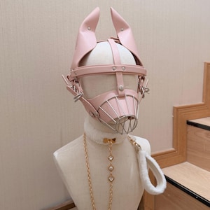 May include: A light pink faux leather headpiece featuring upright pointed ears and a metal wire muzzle cage. The set includes a white faux fur collar with a gold-toned chain leash adorned with three clear crystal-like accents and a white fur handle.