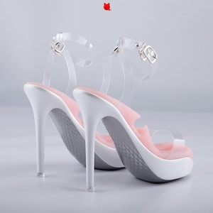 May include: A pair of high-heeled sandals featuring white stiletto heels, a white platform base, and a light pink cushioned footbed. The shoes include clear transparent ankle straps with silver-toned metal buckles and a clear strap across the toe.