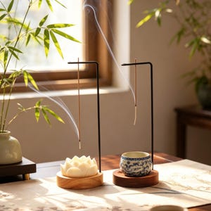 Può includere: Due porta incenso in metallo nero con bastoncini di incenso che bruciano. Uno ha una base a forma di fiore di loto bianco, l'altro una piccola tazza con motivi blu e bianchi. Entrambi sono su basi di legno, con fumo che sale.