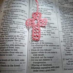 May include: A pink, handmade, crocheted cross bookmark lies on an open book. The cross is made of delicate, openwork crochet and hangs from a matching pink cord. The book's pages display printed text.