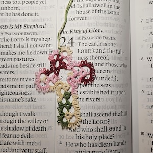 May include: A handmade, colorful crochet cross bookmark with a green, cream, pink, and burgundy floral design. The bookmark is placed on an open book with text visible. The bookmark is a religious item.
