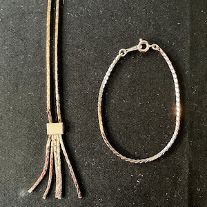 Könnte beinhalten: Eine silberfarbene Halskette mit quadratischem Anhänger und mehreren herabhängenden Strängen, kombiniert mit einem passenden Armband. Der Schmuck wird auf einer schwarzen Oberfläche präsentiert, um seine reflektierenden Eigenschaften und sein detailliertes Design hervorzuheben.