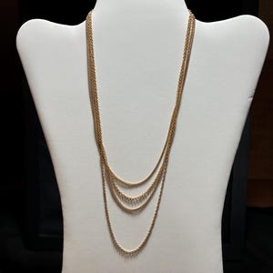 May include: A multi-strand gold-tone necklace featuring four delicate chains of varying lengths that drape in a layered fashion. The chains are constructed with fine, interlocking links that create a consistent, shimmering texture throughout the piece.