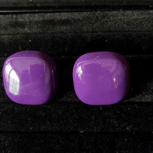 May include: Two glossy, square-shaped, purple earrings. The earrings are a vibrant shade of purple and have a smooth, rounded surface. They are displayed on a black velvet surface, creating a contrast that highlights their color and shape.