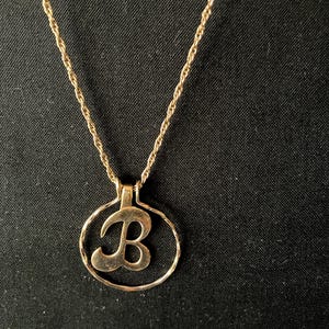 Puede incluir: Un collar dorado con un colgante con la letra "B" dentro de un marco circular. La cadena tiene un diseño de cuerda trenzada, sobre un fondo de tela oscura.