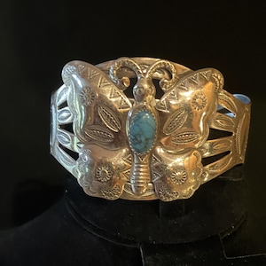 May include: A silver-toned cuff bracelet with a butterfly design. The butterfly has detailed wings and a turquoise stone in the center. The bracelet is displayed on a black surface.