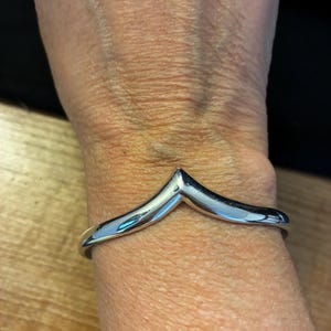 May include: A polished silver-tone metal cuff bracelet featuring a distinct V-shaped chevron design at the centre. The bracelet has a smooth, reflective surface and a continuous band that wraps around the wrist, showcasing a clean and simple aesthetic.