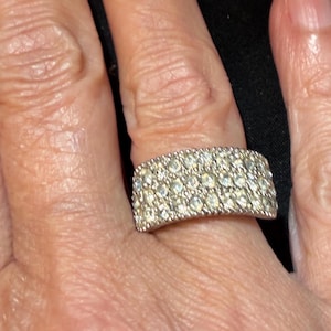 Op de afbeelding: Een brede zilverkleurige ring met drie horizontale rijen kleine, heldere, rond geslepen stenen die dicht op elkaar zijn gezet over het gehele oppervlak van de metalen band, wat zorgt voor een sprankelend uiterlijk op de vinger.