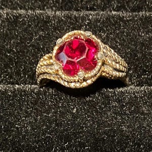 Könnte beinhalten: Ein goldfarbener Ring mit einem großen, facettierten, roten Edelstein in der Mitte. Das Band des Rings hat ein geflochtenes Design, und der Edelstein ist von kleineren, klaren Steinen umgeben. Der Ring ist vor einem dunklen, strukturierten Hintergrund platziert.