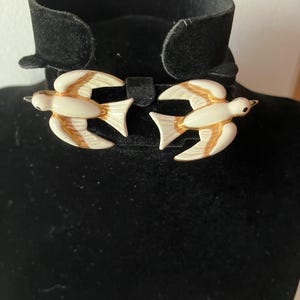 May include: A pair of vintage bird brooches. Each brooch features a white bird with tan and gold accents. The birds are attached to a black velvet collar. The brooches are likely made of enamel and metal.