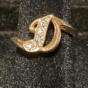 May include: A gold-colored ring featuring the letter "D" embellished with small, clear gemstones. The ring has a delicate, swirling design and is set against a dark, textured background.