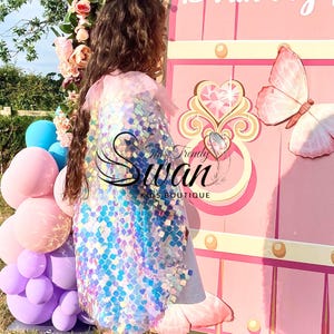 Könnte beinhalten: Ein Umhang, der mit schillernden, quadratischen Pailletten bedeckt ist, die verschiedene Farben reflektieren. Das Kleidungsstück hat einen hellrosa Tüllkragen am Ausschnitt. Der Text The Trendy Swan KIDS BOUTIQUE ist mittig in schwarzer Schrift gedruckt.