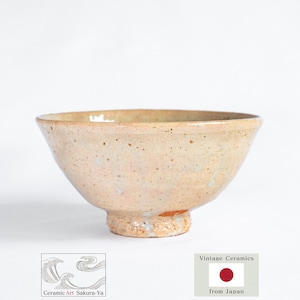 Op de afbeelding: Een beige keramische kom met een brede, schuine rand en een kleine, getextureerde basis. De kom heeft een gespikkelde uitstraling en een enigszins ruwe textuur. De woorden "Ceramic Art Sakura-Ya" en "Vintage Ceramics from Japan" zijn zichtbaar.