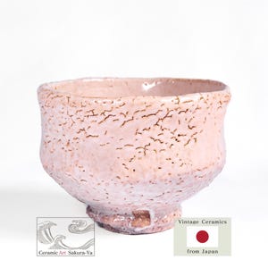 May include: A light pink ceramic tea bowl with a textured, crackled glaze finish. The bowl features a wide, rounded rim and a tapered base. Two small rectangular logos are visible at the bottom, one with wave illustrations and text, the other with a red circle.