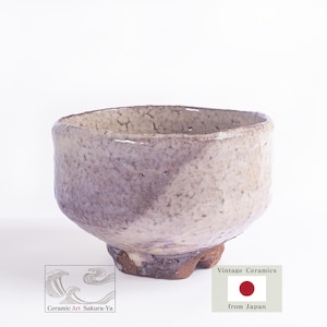 May include: A ceramic tea bowl with a crackled, off-white glaze and a subtle purple-grey hue. The bowl features an uneven, organic rim and a dark, textured base. The text 'Ceramic Art Sakura-Ya' and 'Vintage Ceramics from Japan' is visible below.
