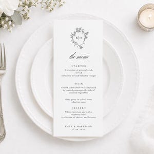 May include: A wedding menu card featuring a floral wreath design and the initials "KH". The menu details a starter, main course, and dessert. The card is on a white plate with a fork and a lit candle in the background.