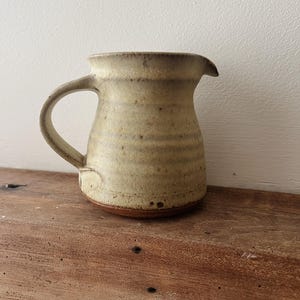 May include: A handmade ceramic jug with a rounded body, a curved handle, and a spout. The jug has a speckled, beige glaze with brown accents and sits on a wooden surface. The jug is approximately 15 cm tall.