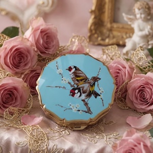 Könnte beinhalten: Ein runder Kompaktspiegel mit gewelltem Rand und hellblauer Emaille-Oberfläche. Der Deckel zeigt eine detaillierte Illustration von zwei Stieglitzen auf einem blühenden Zweig mit weißen Blüten und braunen Stielen, eingefasst in einen goldenen Rand.