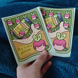 Op de afbeelding: Twee stickers met een cartoon illustratie van een appel spritzer en een roze vrucht met een groen blad. De tekst "My little pokepop" is geschreven in een handschrift stijl. De stickers worden tegen een blauwe achtergrond gehouden.