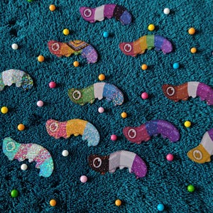 May include: A selection of colourful, glittery, worm-shaped pins scattered on a teal textured surface. The pins feature various colour combinations, including rainbow, pink, and purple. Small, colourful, round sweets are also scattered around the pins.