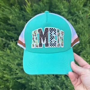 May include: A bright turquoise trucker hat featuring a white mesh back and a central patch with the word AMEN. Each letter has a distinct pattern, including camouflage, leopard print, and black and white checkerboard. The sides feature pink and brown stripes.