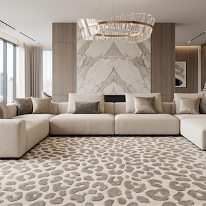 Op de afbeelding: Een moderne woonkamer met een grote, crèmekleurige hoekbank en meerdere kussens. Een patroonkleed met een luipaardprint bedekt de vloer. Boven de bank hangt een decoratieve kroonluchter.
