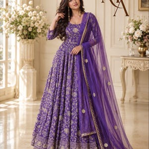 May include: A floor-length purple gown with long sleeves and a round neckline, featuring intricate gold floral embroidery throughout. The dress is paired with a matching sheer purple dupatta with gold-trimmed edges and small floral motifs.