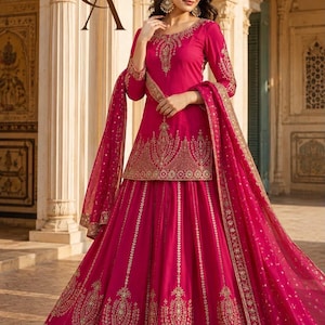 May include: A vibrant fuchsia lehenga choli set featuring a long-sleeved tunic with intricate gold embroidery around the neckline and hem, paired with a matching pleated floor-length skirt and a sheer dupatta, both adorned with gold floral motifs.
