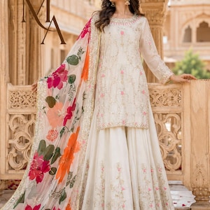 May include: A cream-colored long-sleeved tunic and matching floor-length skirt featuring intricate gold embroidery and pink floral accents. A sheer, lightweight shawl with a vibrant orange and pink floral print drapes over the shoulder.