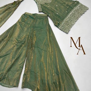 Op de afbeelding: Een saliegroene mouwloze top met zilverkleurig borduurwerk en een geschulpte zoom, gecombineerd met een wijde broek van bijpassende stof met een metallic glans en een bijpassende dupatta. De letters MA staan in bruin rechts afgebeeld.