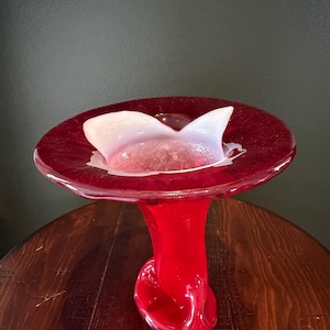 Puede incluir: Un florero de vidrio rojo vibrante con un borde circular ancho y plano y una base escultural retorcida. El centro del florero contiene una pieza insertada translúcida de color rosa pálido en forma de pétalo que contrasta con el rojo.
