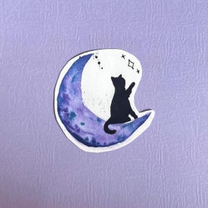 Könnte beinhalten: Aquarell-Illustration einer schwarzen Katze auf einem Halbmond. Der Mond ist in Lila- und Blautönen gemalt, wobei die Katze nach Sternen greift. Der Hintergrund ist helllila.