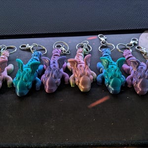 May include: A collection of colorful, 3D-printed dragon keychains. Each keychain features a detailed dragon design in shades of teal, pink, purple, and blue, attached to a silver keyring. The keychains are arranged in a row.