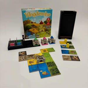 Op de afbeelding: Bordspel "Kingdomino" met een doos met een kasteelafbeelding. Het spel bevat miniatuur kasteelstukken, kleurrijke tegels en een zwarte spelstukhouder. Het spel is op een wit oppervlak geplaatst.
