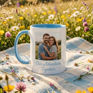 May include: A white ceramic mug with a light blue interior and handle, featuring a printed photograph of a couple embracing and the text "Our Adventure Together" in a blue script font, positioned on a blue and white checkered fabric.