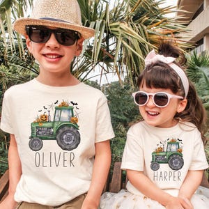 May include: Two cream-coloured short-sleeve t-shirts featuring a green tractor graphic with orange jack-o'-lanterns, black bats, and spiderwebs. The left shirt has the name OLIVER printed below, and the right shirt has the name HARPER printed below.