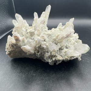 May include: A cluster of clear, pointed quartz crystals emerging from a textured, light gray rocky matrix. The mineral specimen features multiple sharp, translucent crystal points of varying sizes extending outward from a central, rugged base.