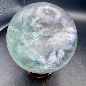 May include: A spherical, translucent mineral orb featuring internal cloudy inclusions and subtle green and purple color variations. The polished stone rests upon a small, dark reddish-brown wooden stand, showcasing a smooth, reflective surface.