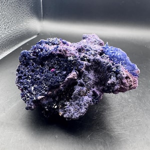 May include: A raw mineral specimen featuring a cluster of deep blue and vibrant purple crystalline structures. The surface is highly textured with jagged, porous formations and small, reflective facets that catch the light across the irregular shape.