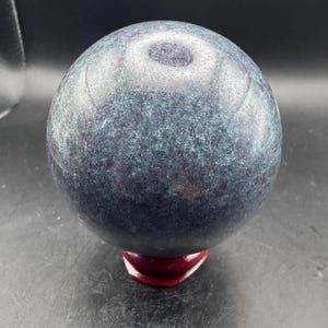 May include: A polished spherical stone with a mottled dark blue and grey surface featuring subtle deep red inclusions throughout the mineral structure, resting on a small, smooth, dark reddish-brown circular wooden display stand.