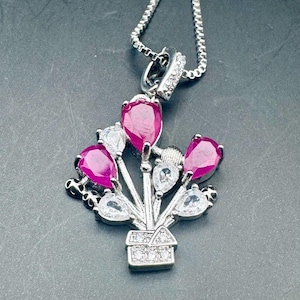 May include: A silver pendant necklace with a bouquet design. The pendant features five pear-shaped, deep pink gemstones and four clear, teardrop-shaped stones. The bouquet is set in a small, square, gem-studded vase, and hangs from a silver chain.