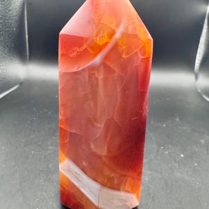 May include: A polished carnelian crystal tower with a hexagonal base and a pointed top. The stone features a vibrant gradient of translucent orange, deep red, and burnt amber hues, accented by distinct, opaque white mineral banding throughout.