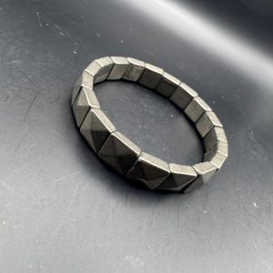 May include: A silver-toned bracelet with a geometric design. The bracelet is made of square-shaped segments with a pyramid-like texture. The bracelet is designed to stretch and fit a variety of wrist sizes. The jewelry is a fashion accessory.