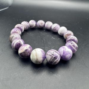 May include: A beaded bracelet featuring large, round beads in shades of purple and white. The beads have a marbled appearance, with swirling patterns. The bracelet is strung on a purple elastic cord, and is a piece of jewelry.