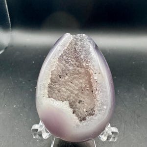 May include: An egg-shaped amethyst geode with a smooth, polished light purple exterior and a central cavity filled with sparkling, translucent crystalline structures. The geode is supported by a clear, transparent plastic display stand.